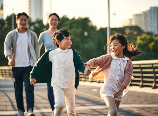 A family crossing a wide pedestrian bridge as the sun begins to set. The two children are laughing and running ahead of their parents, who are walking behind holding hands while smiling.