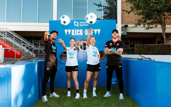 Four soccer players pose in front of a blue 'Blacktown Quarter Kick-Off' goal display outdoors.