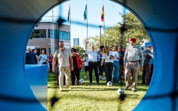 People walk toward a soccer goal game on the lawn, viewed through a circular opening in the goal display.