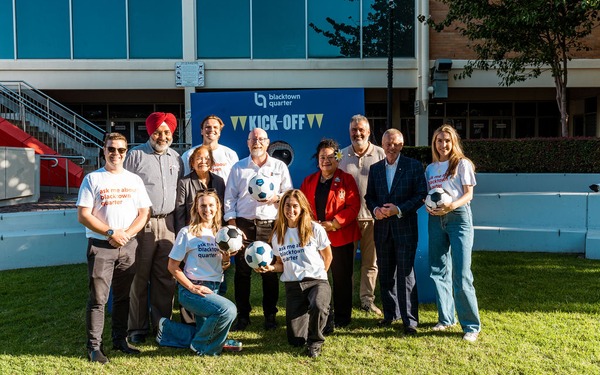 Group photo of event participants and staff posing with soccer balls in front of a Blacktown Quarter Kick-Off display.