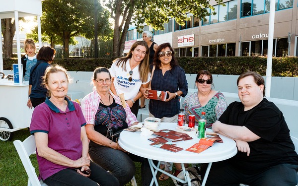 Group of community members seated around a table with event staff standing behind them at an outdoor gathering.