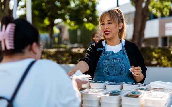 A woman serves takeaway food containers from a stall at an outdoor community event.
