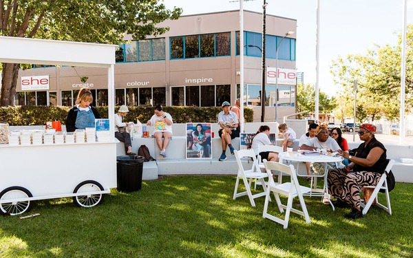 People sit and chat on white chairs beside a small stall during a community gathering on a grassy plaza.