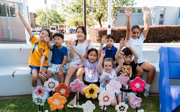 Group of children smiling and waving behind a display of colorful crocheted flowers at an outdoor event.
