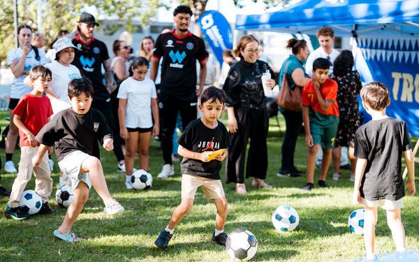 Children play with soccer balls on the grass while adults and players stand nearby at a community event.